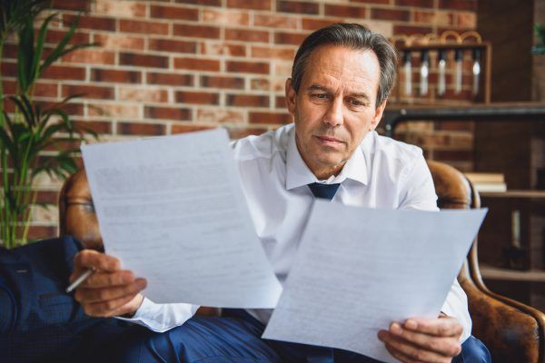 Exhausted mature businessman is sitting in room. He holding documents and reading it