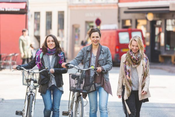 Group of women walking in Copenhagen. They are in their twenties and they are wearing smart casual clothes. Two of them are holding a bicycle, typical mode of transport in Denmark.