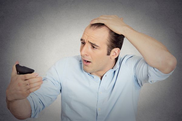 Closeup portrait shocked man feeling head, surprised he is losing hair, receding hairline or seeing bad news on cell phone isolated grey wall background. Negative facial expressions, emotion feeling