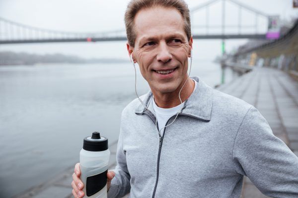 Close up runner in gray sportswear standing near the river with bottle of water in hand and looking away
