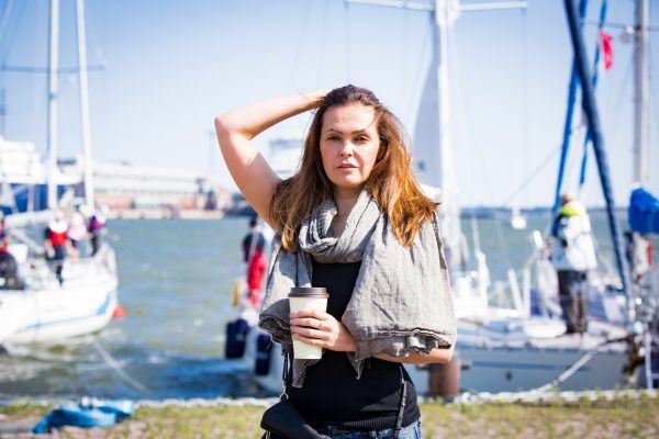 Portrait of smiling stylish middle aged woman walking along sea bay. Sailboats in harbour, seaside. Sunny summer day. Wind in the hair, drinking coffee. Vacation destination. Helsinki. Finland