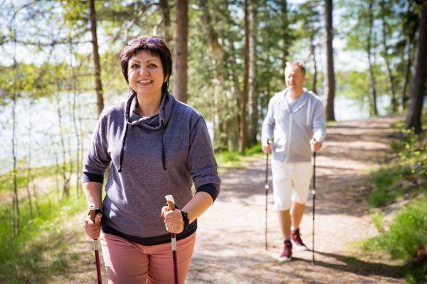 Summer sport in Finland - nordic walking. Man and mature woman hiking in green sunny forest. Active people outdoors. Scenic peaceful Finnish summer landscape.