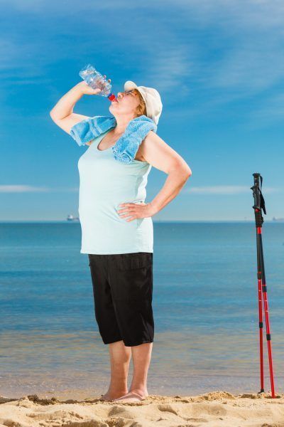 Senior woman practicing nordic walking on sea shore, drinking fresh water after activity. Elderly female enjoying sunny summer day. Healthy active retirement age.